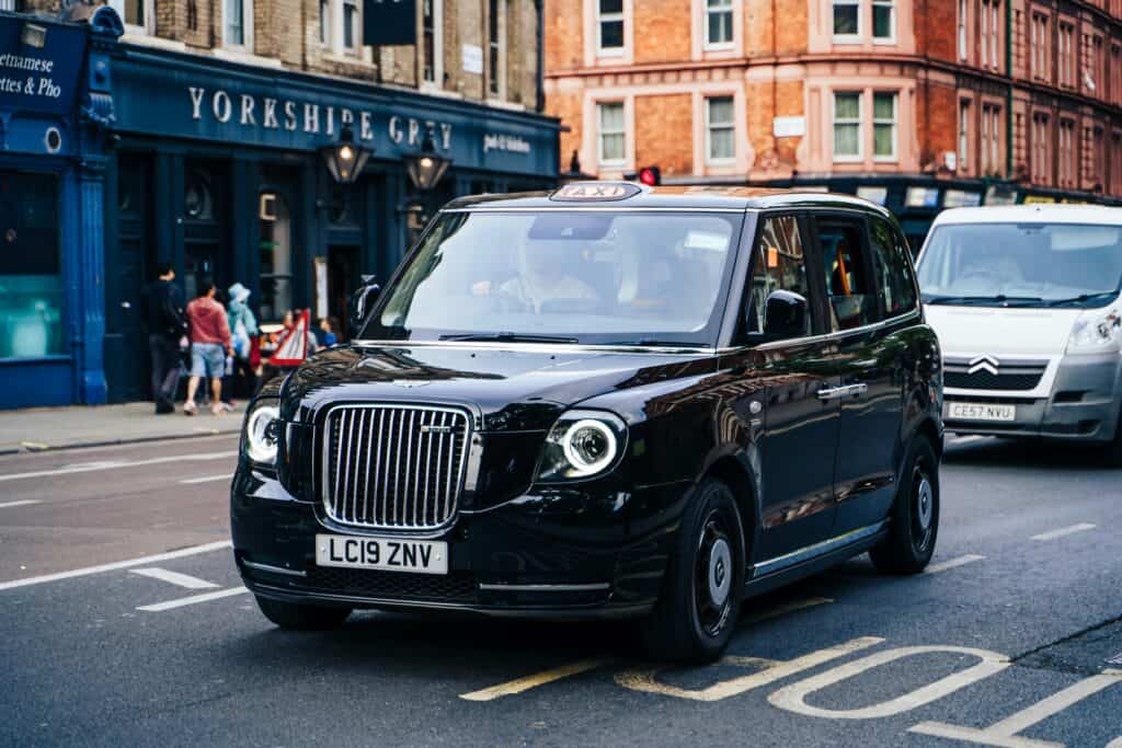 A classic London Black Cab on a high-street city road during the daytime.