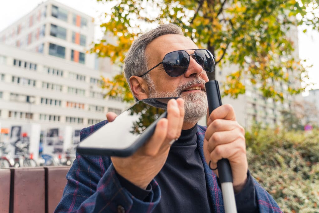 A blind middle-aged man with gray hair and beard wearing sunglasses holding cane and phone using text-to-speech function.