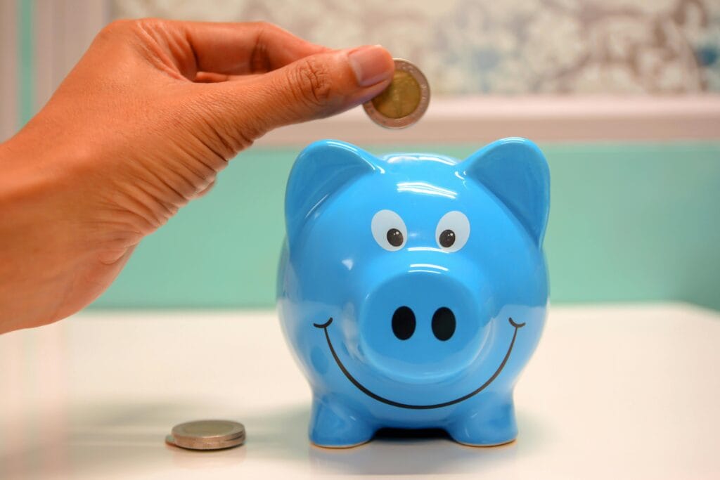 A close up photo of a hand placing a Euro coin into a turquoise blue piggy bank. The face of the bank has a smiling pig's face.