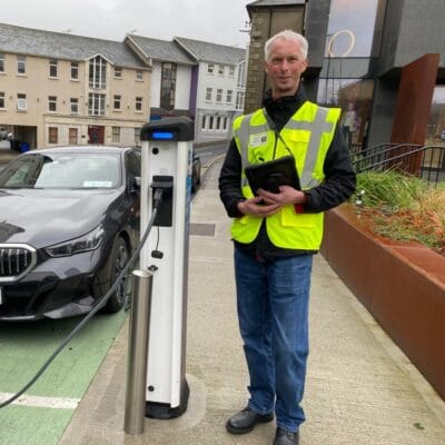 A Caucasian man and access consultant, Nick Holland, wearing a hi-vis jacket and holding a tablet smiles and poses for a photograph on a sidewalk in Wexford, Ireland. He is stood next to an Electric Vehicle charging point.