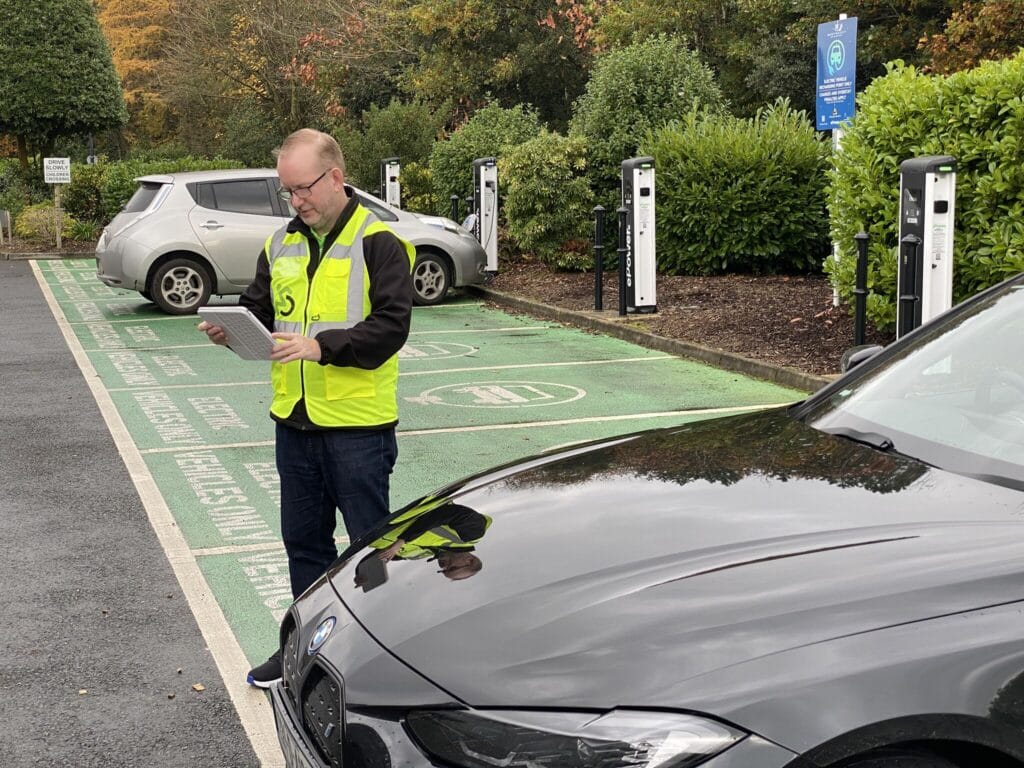 Direct Access’ Steve Dering in high vis jacket with tablet. Two electric cars are parked in a ten bay charging station.