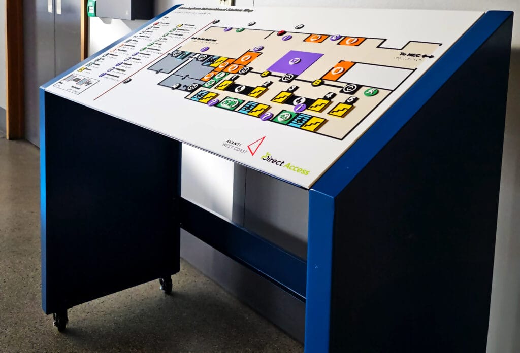 A wide shot photograph of the new Birmingham International tactile braille map board shot using deep focus. The face of the map board provides a colourful rendition of Coventry Station's layout using a raised braille key and symbols against a white background. In the bottom left corner QR codes are available for audio described and sign language elements.