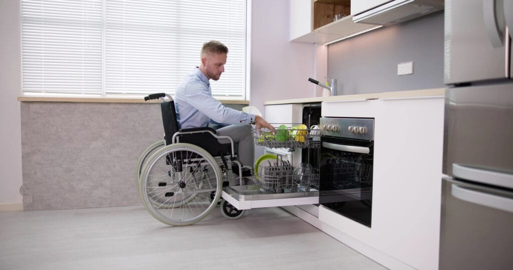 A young Caucasian man in a wheelchair takes cutlery items out of a split-height counter dishwasher in his home kitchen.