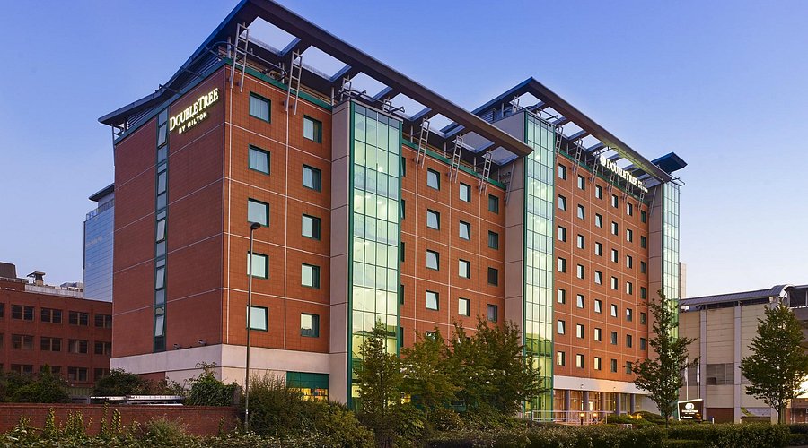 Modern DoubleTree by Hilton hotel building with a red-brick façade and vertical glass sections, photographed at dusk with soft blue sky and landscaped greenery in the foreground; the hotel name is visible at the top of the building.