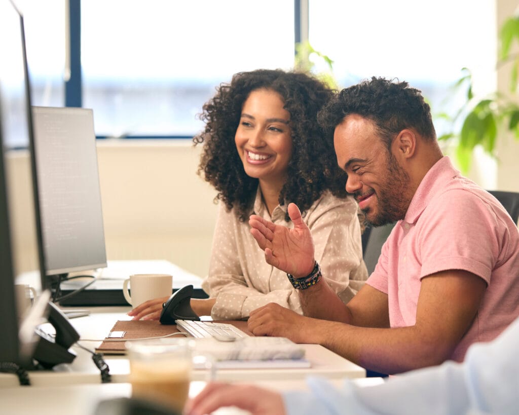 An image of a mixed-race woman working in an office environment, she is smiling at a computer screen on a desk with a coffee cup, a keyboard, and two monitors. Next to her is a man she is training, who has down syndrome and is also smiling.