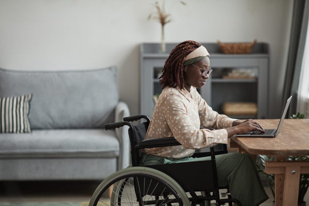 Side view portrait of young African-American woman using wheelchair while working from home on a laptop in a minimal grey living room interior.