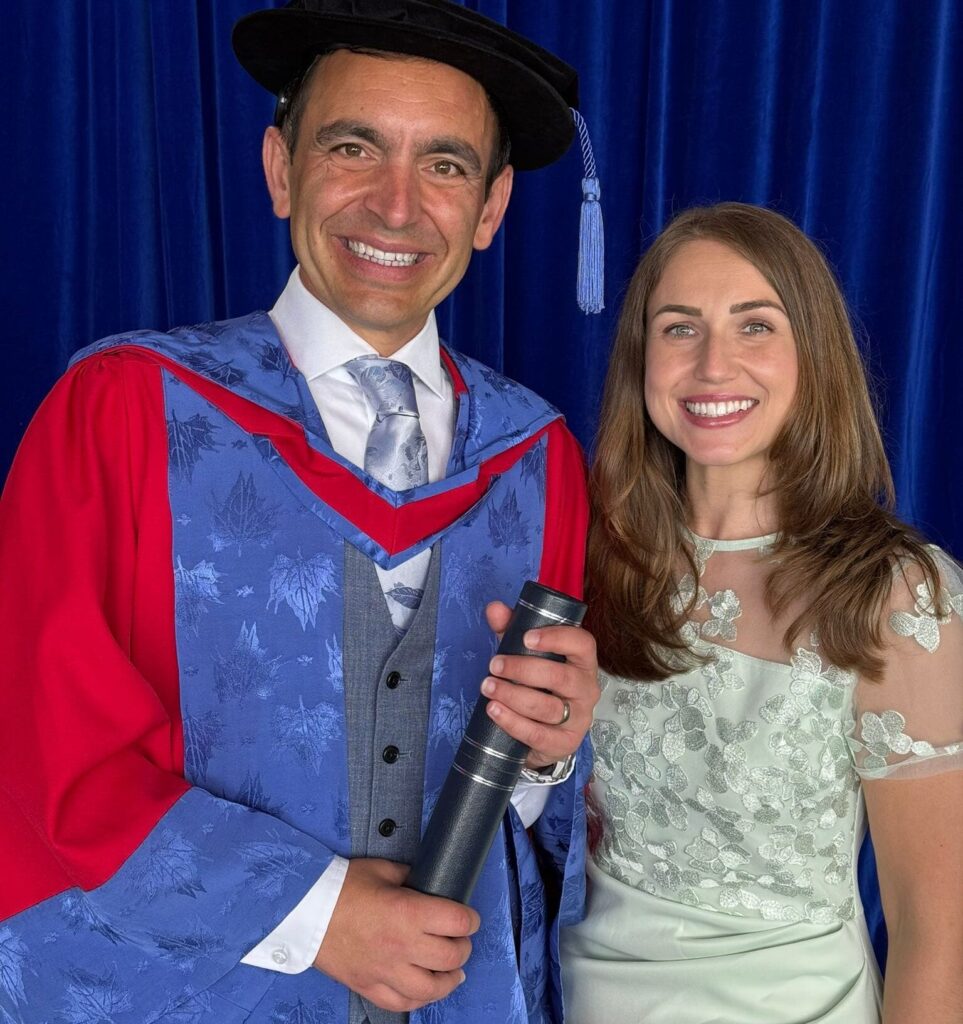 A photograph of Steven Mifsud MBE posing for a photo alongside his wife, Direct Access Co-Director Jude Mifsud. Steven is wearing a blue and red graduation robe and holding a black tubular scroll containing his doctorate. Jude is wearing a half-sequined silver dress. Both are smiling.