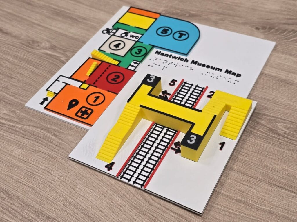 Image description: A close-up of a 3D-printed tactile model designed for accessibility or educational use. The model features a yellow structure with black borders and staircases on both sides, positioned on a white base. A black platform labeled with a raised white number "3" sits centrally, while black and red tactile lines resembling train tracks run across the base. Raised black numbers "1" and "4" are also visible, suggesting a layout of train platforms or station zones intended to aid spatial orientation for visually impaired users.