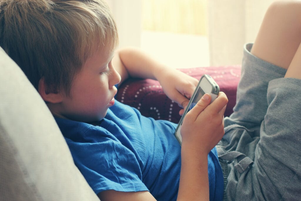 Image description: A young child wearing a blue t-shirt and gray shorts reclines on a couch, holding a smartphone or handheld device with both hands. The child is focused on the screen. Behind them is a maroon cushion and a softly lit curtain, creating a relaxed indoor setting.