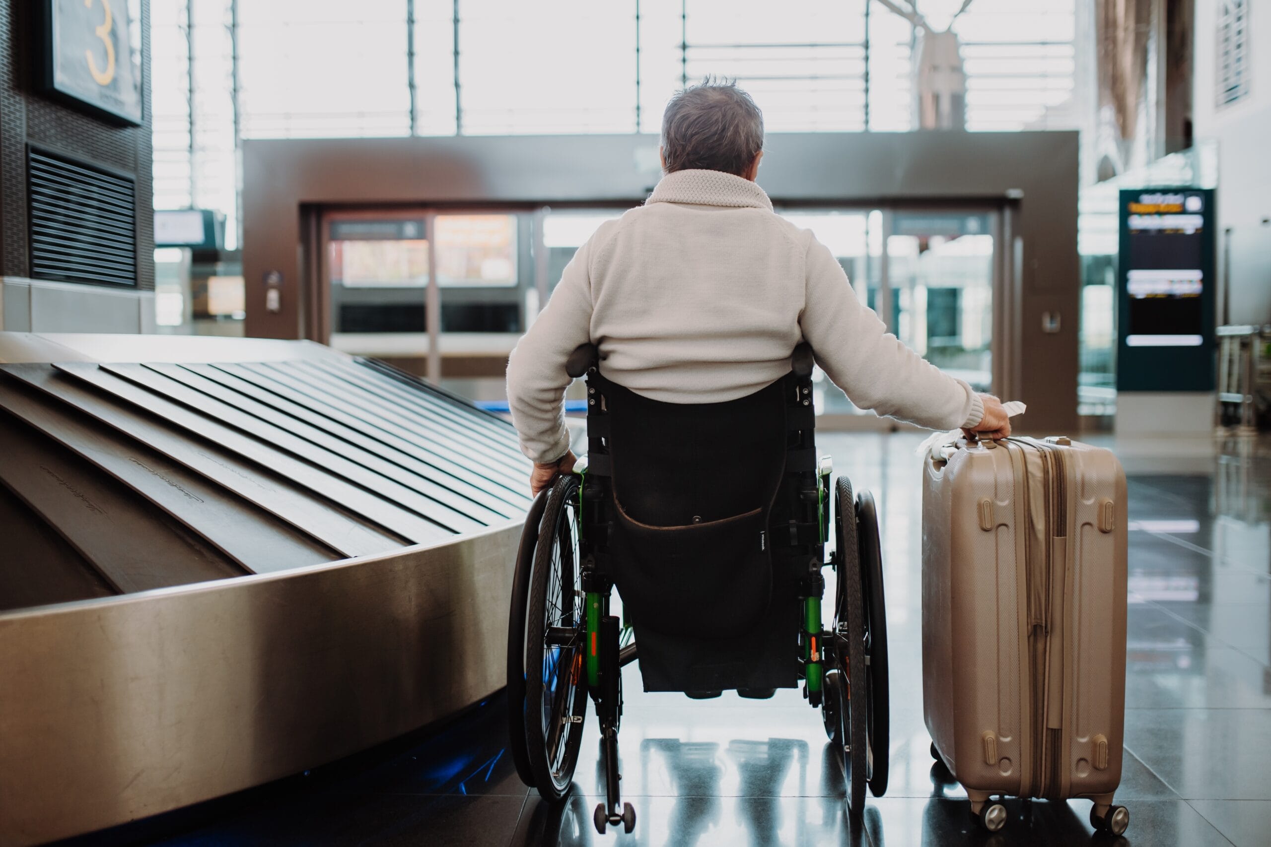 Rear,View,Of,Man,On,Wheelchair,At,Airport,With,His A wide shot photograph of an older white and male wheelchair user with grey hair in an airport foyer. He is sat next to a baggage conveyor belt and has his right arm stretched out, gripping his luggage.