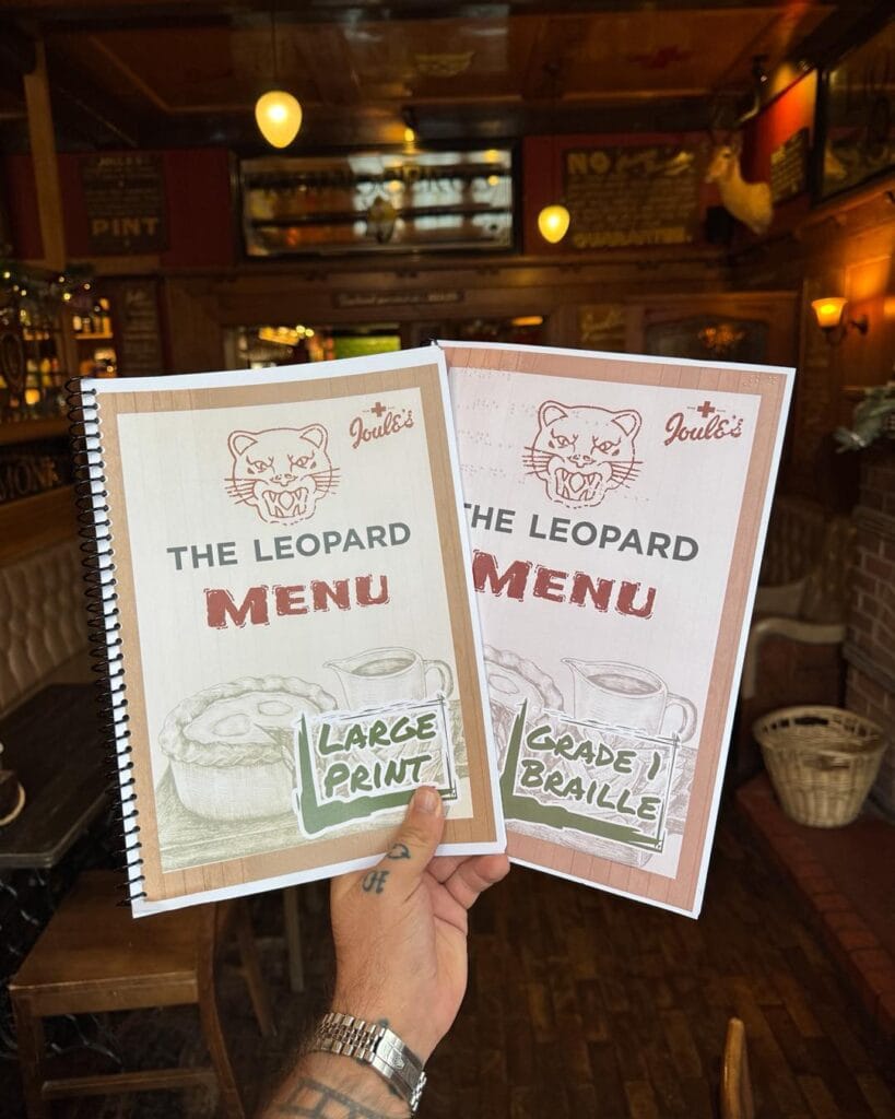 The image shows a hand holding two menu booklets inside a cozy pub or restaurant. The menus are titled “The Leopard Menu” and each features the Joule’s brewery logo at the top. One menu is labeled “Large Print” and the other is labeled “Grade 1 Braille.” Both covers display illustrations of a pie and a drink. The background reveals a warmly lit interior with wooden furniture, vintage-style décor, and hanging lights, creating a classic pub atmosphere.
