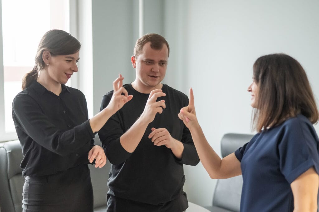 The image shows a mixed group of people standing together in a bright indoor setting, engaged in communication using sign language. All three individuals are positioned in a small circle, facing one another with their hands raised as they form various signs. They are dressed in dark, professional clothing, and their body language suggests focused, attentive interaction. The background features light-colored walls and minimal furniture, giving the scene a calm, classroom‑ or office‑like atmosphere.