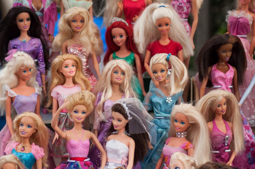 A large collection of fashion dolls arranged closely together in multiple rows. The dolls are dressed in a variety of colorful outfits, including dresses in shades of pink, purple, blue, and red. Many have long, styled hair in different colors such as blonde, brunette, black, and red. Some dolls wear accessories like tiaras, necklaces, and veils, giving the display a vibrant, playful, and decorative appearance. The overall scene looks like a collection or display of dolls, possibly at a market, event, or toy exhibition.