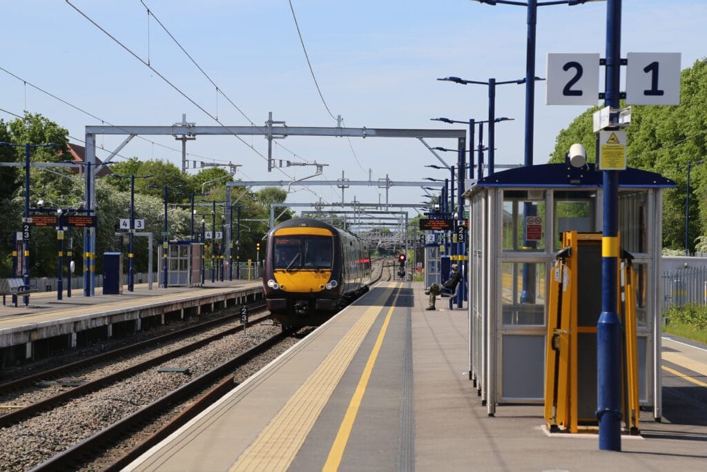 The image shows Bromsgrove railway station with multiple platforms and tracks under bright, sunny weather. A modern yellow‑and‑black train is approaching the station along the center track. The platforms are marked with signs indicating platform numbers, including large overhead signs for platforms 2 and 1 in the foreground. Blue lamp posts and metal structures for overhead electric wires line the tracks. There are small sheltered waiting areas, station signage, and safety markings along the platform edges. Trees and greenery border the station, giving it a clean and open atmosphere.