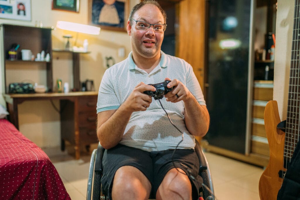 The image shows a caucasian man sitting in a wheelchair inside a cozy, lived‑in room while holding a video game controller. The individual is wearing a light-colored polo shirt and dark shorts, focused on playing a game. Around them, the room features a desk with a lamp, shelves with various items, framed pictures on the wall, and a guitar leaning against furniture. The warm lighting and casual setup create a relaxed, homey atmosphere.