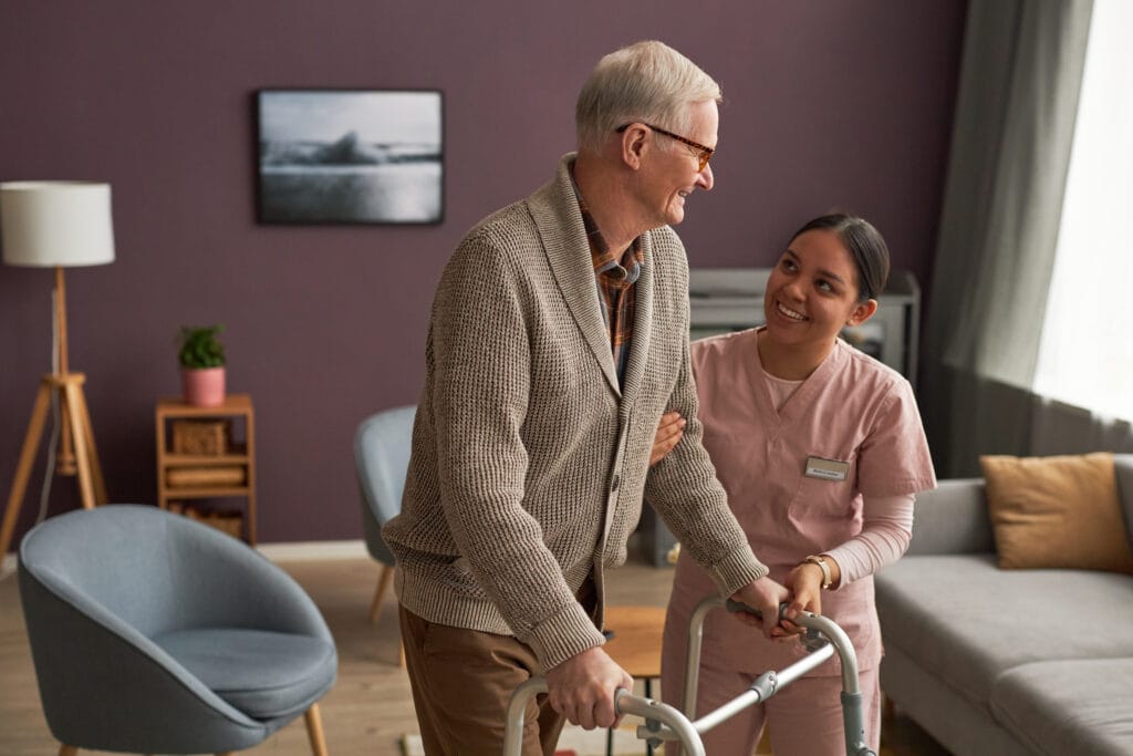 A caregiver in a pink medical uniform supports an older adult who is using a walking frame. They are standing together in a warmly lit living room featuring modern furniture, including a grey armchair, a wooden side table with a potted plant, and a sofa with yellow cushions. A floor lamp and a framed landscape photograph hang on the wall behind them, creating a calm and homelike environment as the caregiver assists the individual with mobility.