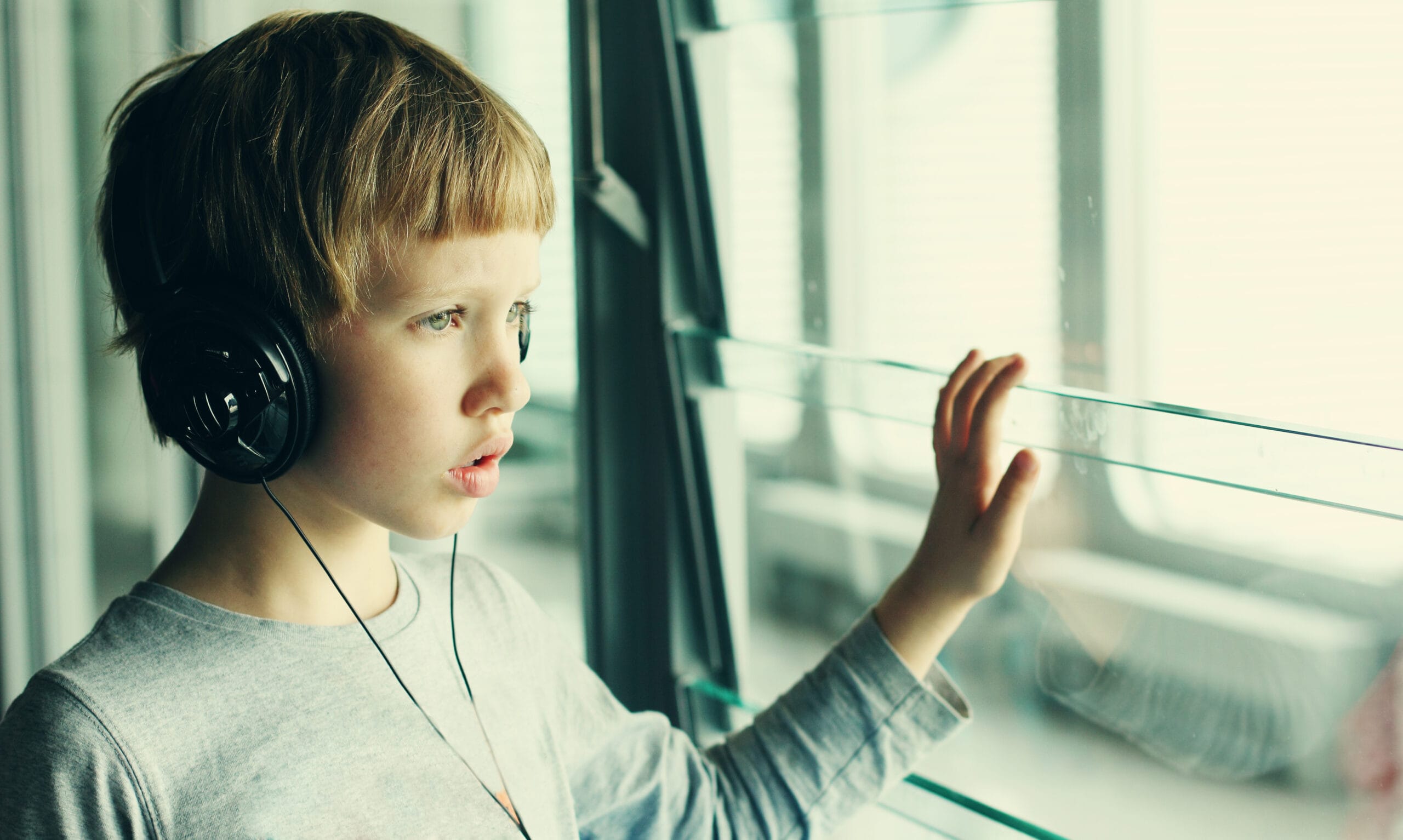 A Caucasian male child standing indoors near a large window, wearing over‑ear headphones. They are dressed in a long‑sleeved, light gray shirt and have one hand gently resting against the glass. Soft natural light comes through the window, creating a calm, reflective atmosphere. The scene suggests someone listening to music or audio while quietly looking outside.