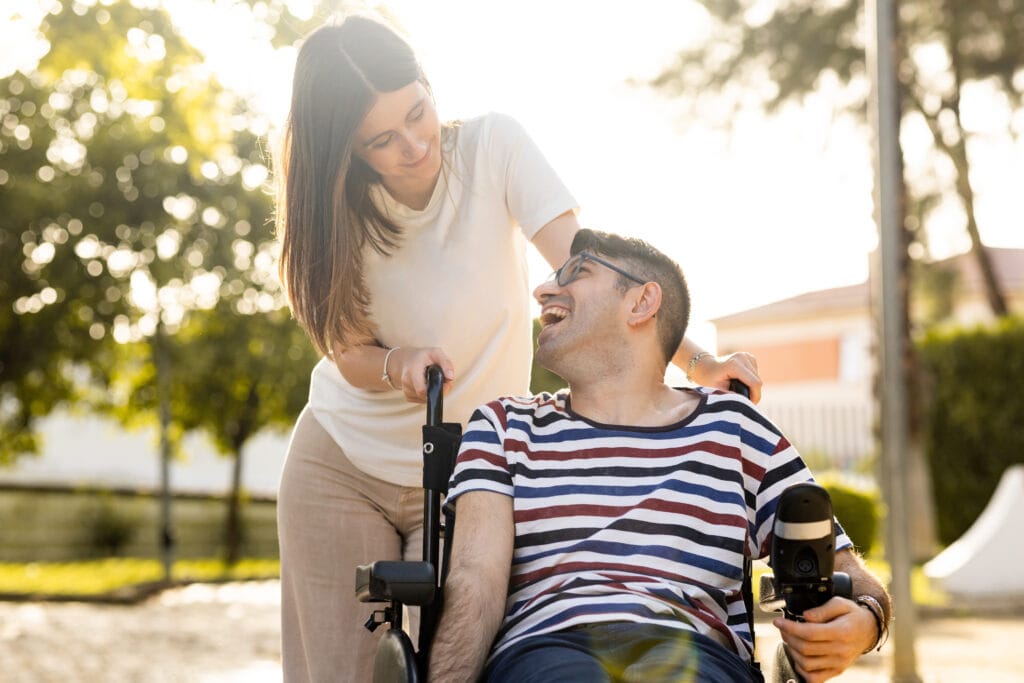 The image shows an outdoor scene with two people interacting in a sunny, park‑like setting. One person is seated in a wheelchair, wearing a striped shirt and holding what appears to be a drink or bottle in one hand. Another person stands beside them, leaning slightly forward while holding the wheelchair’s handle and resting a hand supportively on the seated person’s shoulder. Warm sunlight creates a soft, glowing atmosphere, and trees and buildings are visible in the background, giving the moment a calm and supportive feel.