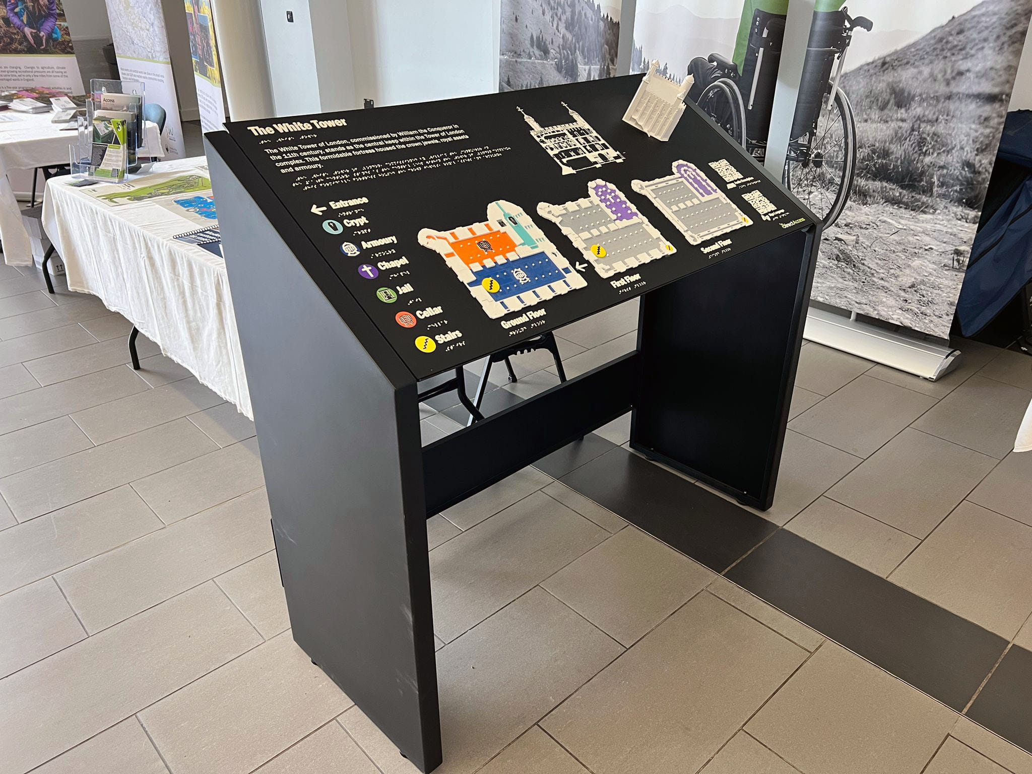 A wide shot photograph of a black tactile braille map board shot in deep focus. The face of the map board provides a colourful rendition of the Tower of London's layout using a white, raised braille key and various symbols against a black background. A large white tactile model of the Tower of London's exerior sits on the face of the right side. Behind the map is a white table and Direct Access display stand.