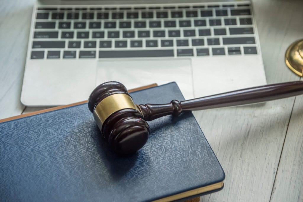 A judge’s gavel made of dark polished wood rests on top of a closed dark blue book, which itself is placed on the keyboard of an open silver laptop. The gavel has a gold band around the head, adding a formal, traditional touch. To the right side of the frame, part of a gold-colored object—likely a scale of justice—is visible but slightly out of focus. Everything sits on a light wood desk, creating a clean, professional setting. The composition visually represents the intersection of law and technology, blending classic legal symbols with modern digital tools.