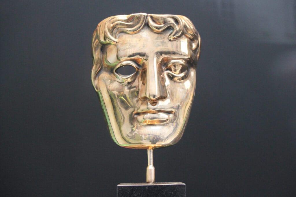 A gold BAFTA trophy in the shape of a stylized human face mask sits on a glossy black rectangular base. The mask features smooth, sculpted facial details—arched brows, a defined nose, closed lips, and wavy hair framing the left side. One eye is hollow, creating a dramatic contrast with the reflective gold surface. The background is softly blurred, keeping the focus on the iconic award associated with excellence in film, television, and games.