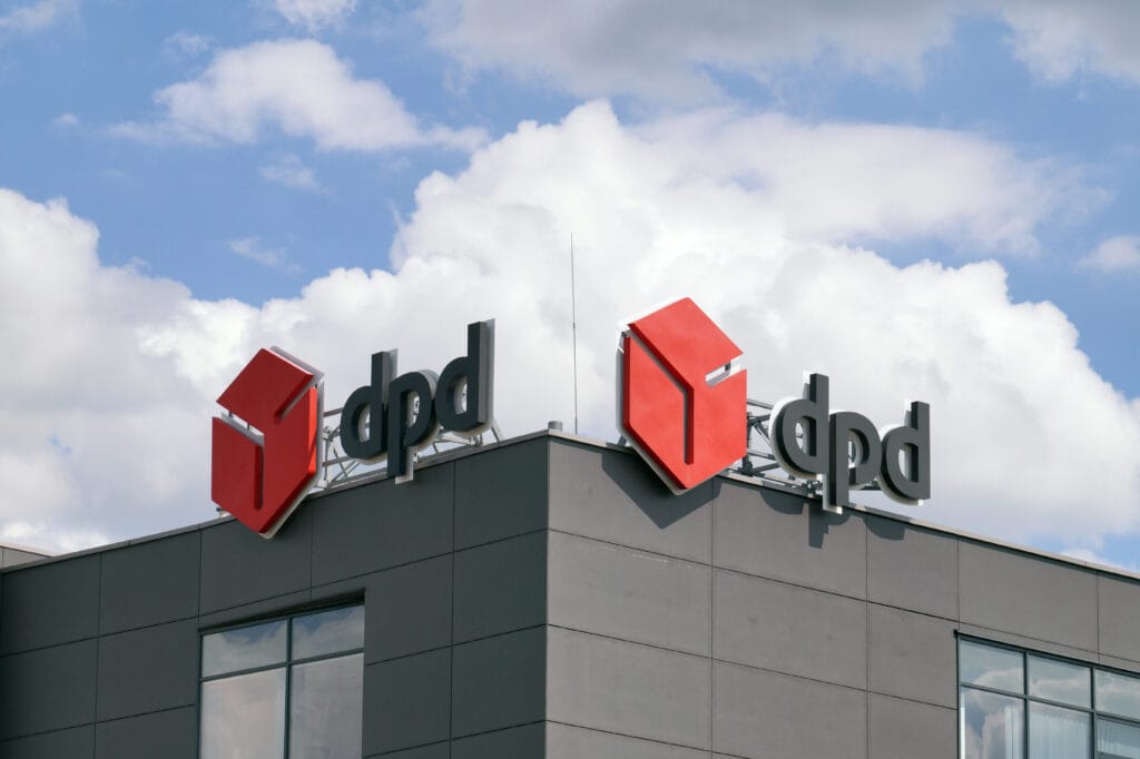 A modern commercial building exterior is shown with two large rooftop signs displaying the dpd logo. Each sign features a bold red cube symbol next to the lowercase letters “dpd” in a clean, black sans‑serif font. The building façade is made of dark gray panels with long horizontal windows running across the upper level. The sky above is partly cloudy, creating a bright but diffused light over the scene. The overall image emphasizes corporate branding and the recognizable identity of the parcel delivery company.