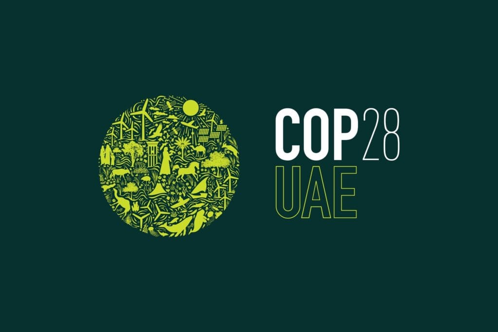 A clean, modern logo for COP28 UAE appears against a plain background. On the left, a circular emblem is filled with small green illustrations representing climate and environmental themes—wind turbines, solar panels, trees, water, animals, and people—arranged like a mosaic to symbolize sustainability and global climate action. To the right of the circle, the text “COP28 UAE” is set in a bold, minimalist font: “COP” in solid white, “28” in a thinner white typeface, and “UAE” outlined in green. The overall design feels contemporary, optimistic, and aligned with environmental advocacy.