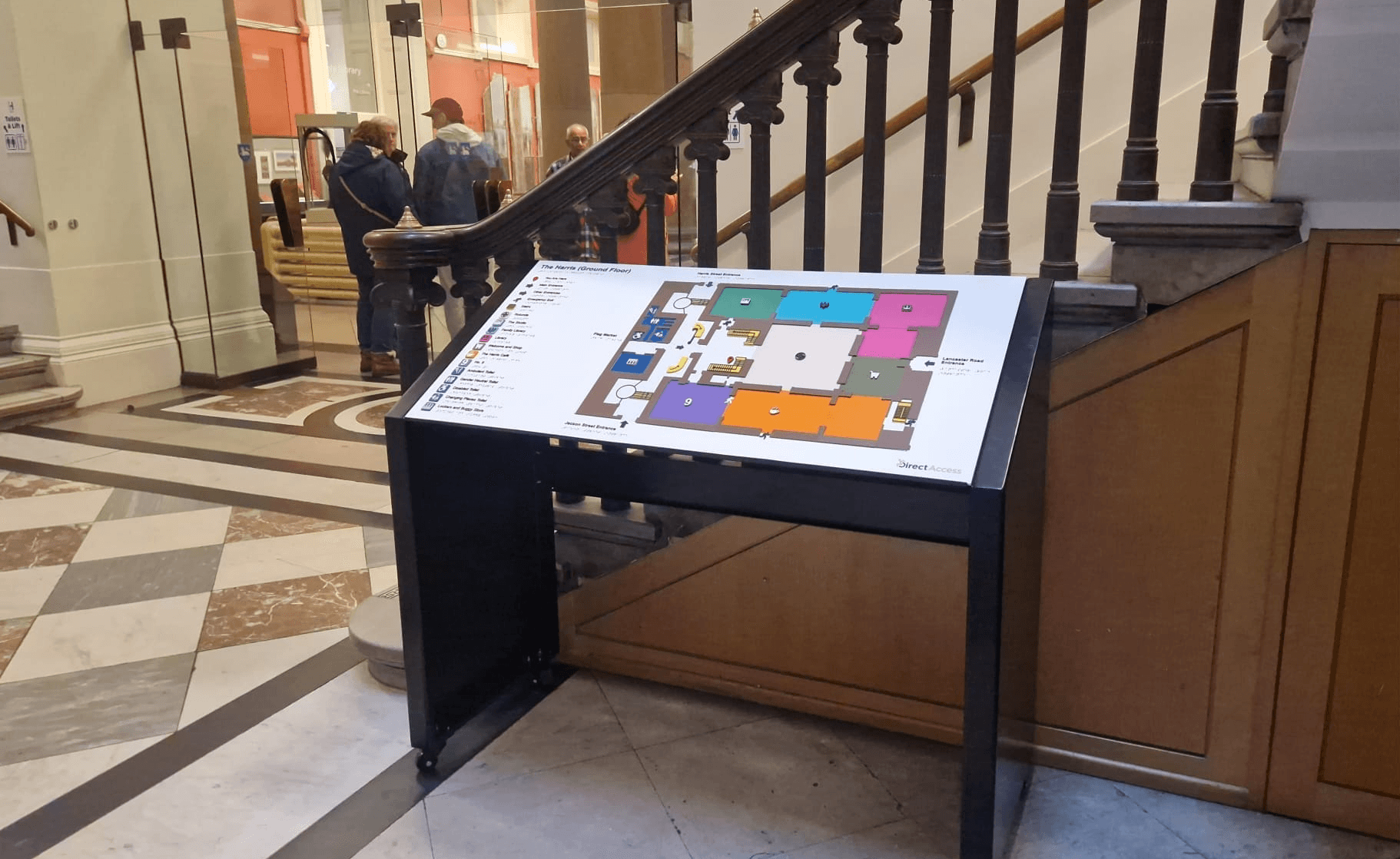 A tactile map on a black stand is positioned near a staircase inside a museum. The map includes raised elements, Braille, and a colour‑coded layout. Behind it is a staircase with wooden railings and a glass wall overlooking the atrium.