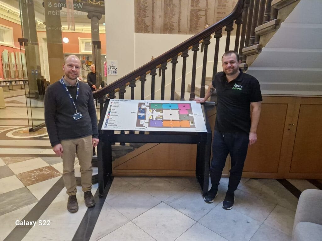 Two people shake hands in a museum atrium. One wears a lanyard; the other wears a black “Direct Access” shirt. They stand in front of a staircase and a large directory map. The space has marble flooring and glass walls.