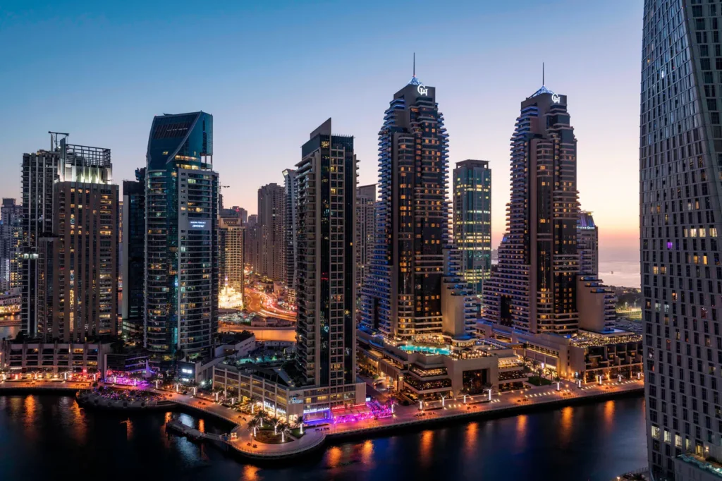 A twilight cityscape featuring a waterfront lined with illuminated high‑rise buildings. Tall modern skyscrapers with bright lights reflect onto the water below, while the sky transitions from deep blue to a warm sunset glow on the horizon.