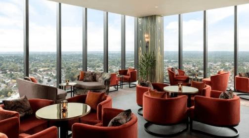 A modern, high‑level lounge with floor‑to‑ceiling windows offering a wide panoramic view of a sprawling cityscape. The room is furnished with circular tables and plush, curved chairs in warm tones of red and orange. Soft lighting, reflective tabletops, and a decorative column with vertical gold accents add to the upscale, relaxed atmosphere.