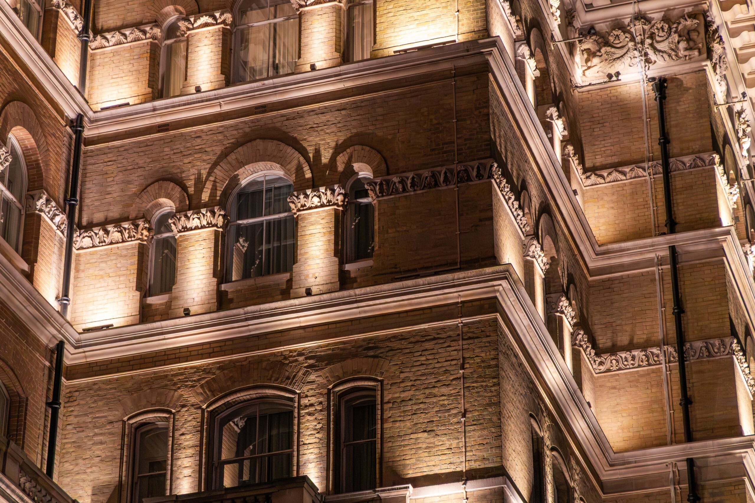 The image shows the exterior of an ornate, multi-story brick building at night, illuminated by warm architectural lighting. The structure features detailed craftsmanship, including arched windows, decorative stone carvings, and layered cornices. Each level of the building has symmetrical window placements, many with rounded tops and framed by intricate brickwork. The corners of the building are emphasized with vertical architectural elements and additional decorative accents, giving it a grand and historic appearance. The lighting highlights the textures of the brick and the elaborate designs, creating a dramatic contrast between light and shadow. Overall, the scene conveys a sense of elegance and classic architecture, possibly resembling a historic hotel or a heritage building in a city setting.
