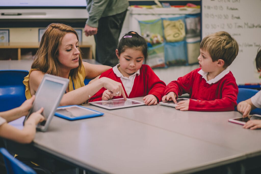 A classroom setting with several young children seated around a rectangular table. An adult is seated with the children at the table. The adult is holding a tablet device and is pointing toward the screen of another tablet placed on the table in front of one of the children. The children are each holding or using tablet devices. The child at the center of the image is looking down toward the tablet on the table. Another child seated to the right is looking toward the adult who is gesturing. Additional children are partially visible around the edges of the table. The table is light-colored, and the chairs surrounding it are blue. Behind the group, there is a whiteboard and various classroom materials, including shelves, posters, and a storage pocket organizer hanging on the wall. The environment appears well-lit, and the focus is on the interaction around the table and the use of digital devices.