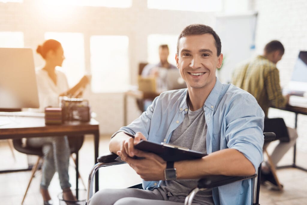 The image shows a smiling caucasian man sitting in a wheelchair in a bright, modern office environment. He appears relaxed and confident, holding a tablet in his hands. He’s wearing a light blue button-up shirt over a gray T-shirt, and a smartwatch on his wrist. In the background, there are a few coworkers engaged in their own tasks—one person sitting at a desk using a computer, and another standing and interacting nearby. The space is well-lit with natural sunlight streaming in, giving the office a warm and collaborative atmosphere. Overall, the scene conveys a professional, inclusive workplace with a positive and productive vibe.