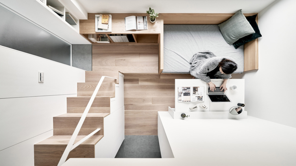 Overhead view of a compact, modern living space with wooden flooring, a small bed and shelving unit, stairs leading to a loft area, and a young Caucasian woman seated at a desk working on a laptop beside neatly arranged items.