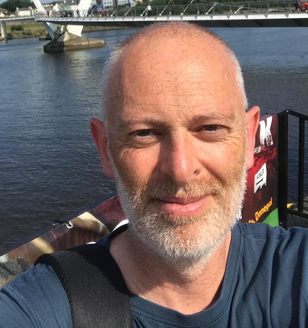 Close-up outdoor selfie of an adult man with a short beard standing near a river, with a modern bridge and buildings in the background.