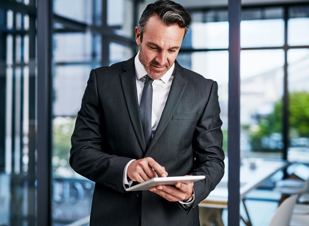 A male business professional in a grey suit standing in a modern office, using a handheld tablet with one finger while large windows and blurred workspace are visible in the background.