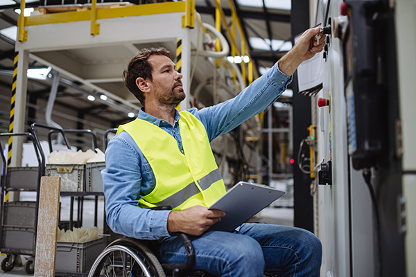 Man in a wheelchair wearing a high-visibility vest uses a control panel in a manufacturing facility while holding a clipboard, with industrial machinery visible in the background.