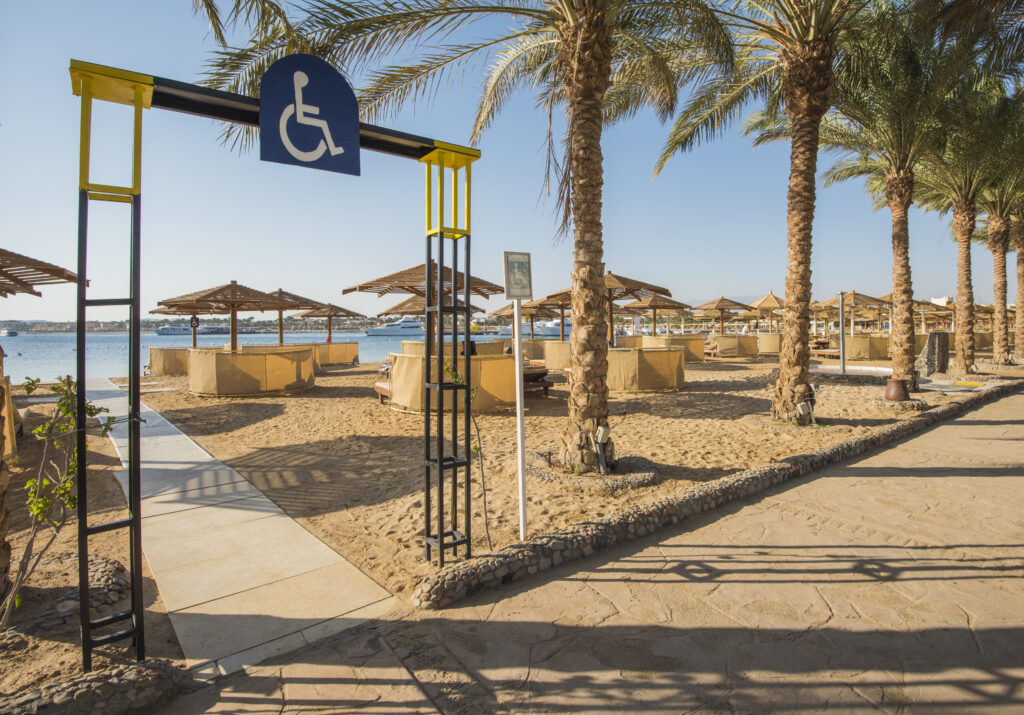 Accessible beach entrance with a wheelchair-accessible walkway leading onto sandy beach areas. A blue accessibility sign is mounted above the pathway. Palm trees line the beach, with shaded seating huts and sun loungers arranged across the sand, and calm water with boats visible in the background under a clear sky.