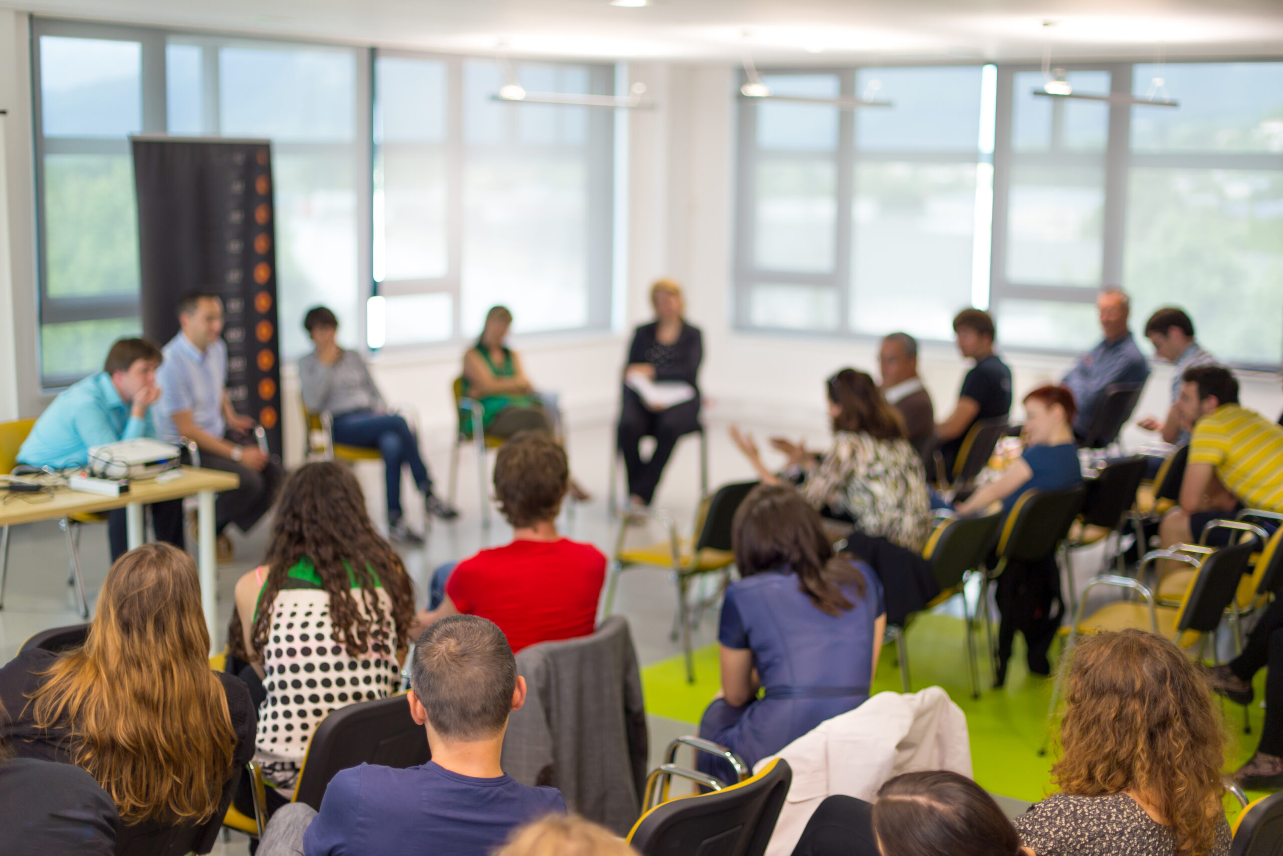 A group of people sit in a circle in a bright meeting room during a workshop or discussion session. Chairs are arranged around the room, with participants listening and speaking. Large windows line the walls, allowing natural light into the space, while a table with equipment is visible on the left. The image is intentionally out of focus, creating a soft, blurred view of the gathering.