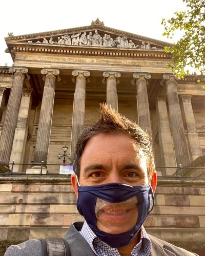 A middle aged Maltese man with short brown hair wearing a clear, see through face mask for lipreading smiles for a selfie in front of the Harris Museum, a building built in a neo-classical design featuring roman styled support pillars. The building is painted a mixture of light and dark brown implying old age and wear.