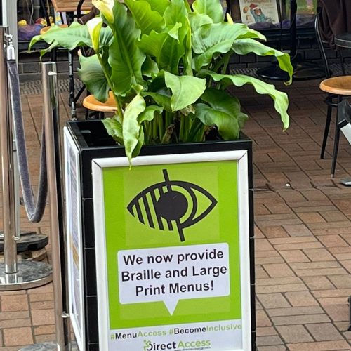 A street sign on an outdoor plant pot that reads "We now provide Braille and Large Print Menus".