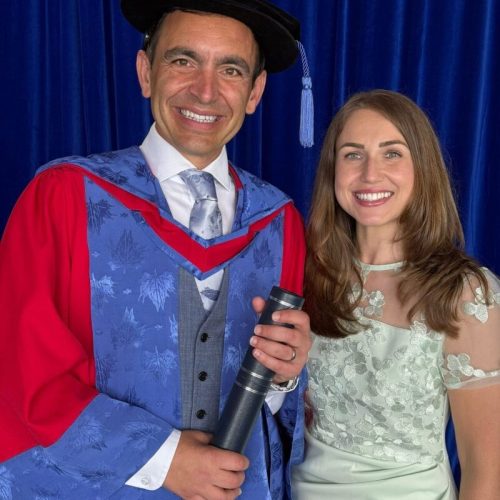 A photograph of Steven Mifsud MBE posing for a photo alongside his wife, Direct Access Co-Director Jude Mifsud. Steven is wearing a blue and red graduation robe and holding a black tubular scroll containing his doctorate. Jude is wearing a half-sequined silver dress. Both are smiling.