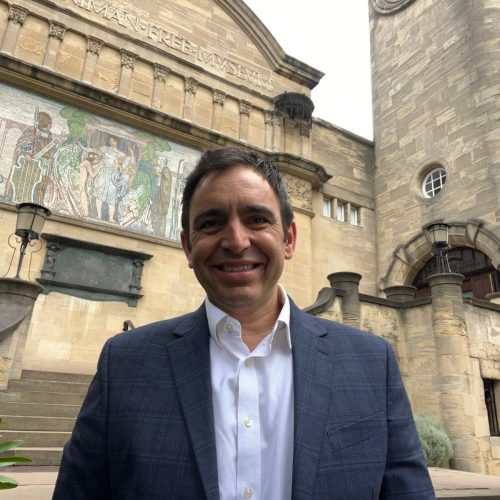 Direct Access founder Steven Mifsud MBE, a Maltese man with short brown hair and a grey suit smiles for a photo in the courtyard of the Horniman Museum and Gardens on an overcast day. Behind him is a stone staircase leading up to a side-entrance of the museum.