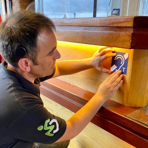 A man places a Hearing Loop sticker onto the underside of a desk.