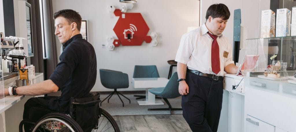 Two male disabled co-workers; one a male wheelchair user, the other a man with down syndrome in a kitchenette. The former is using a coffee machine, the latter watching a video on a tablet.
