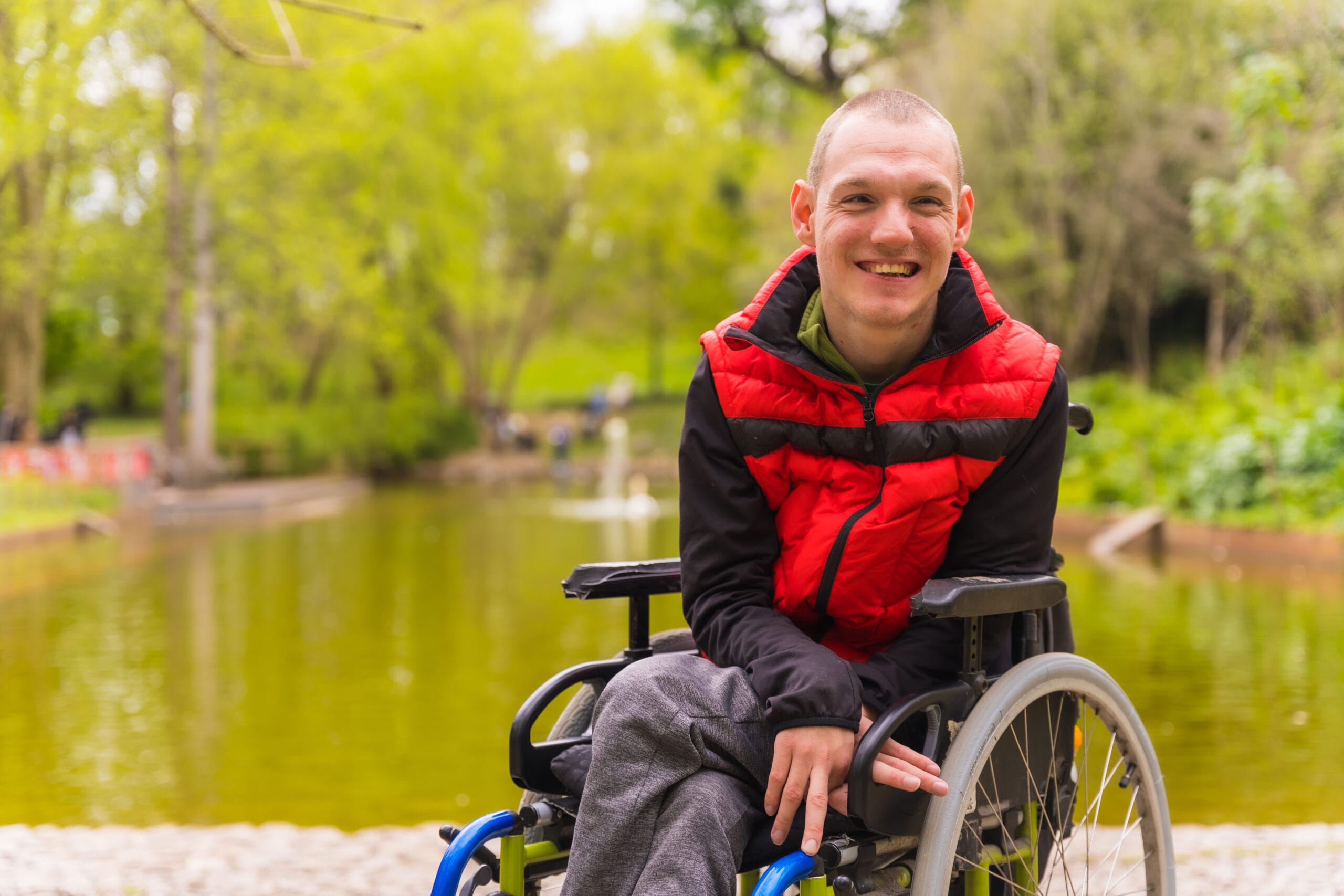 Portrait of a young man with learning disabilities in a wheelchair in a public park smiling. Behind him is a small lake and a sea of trees.