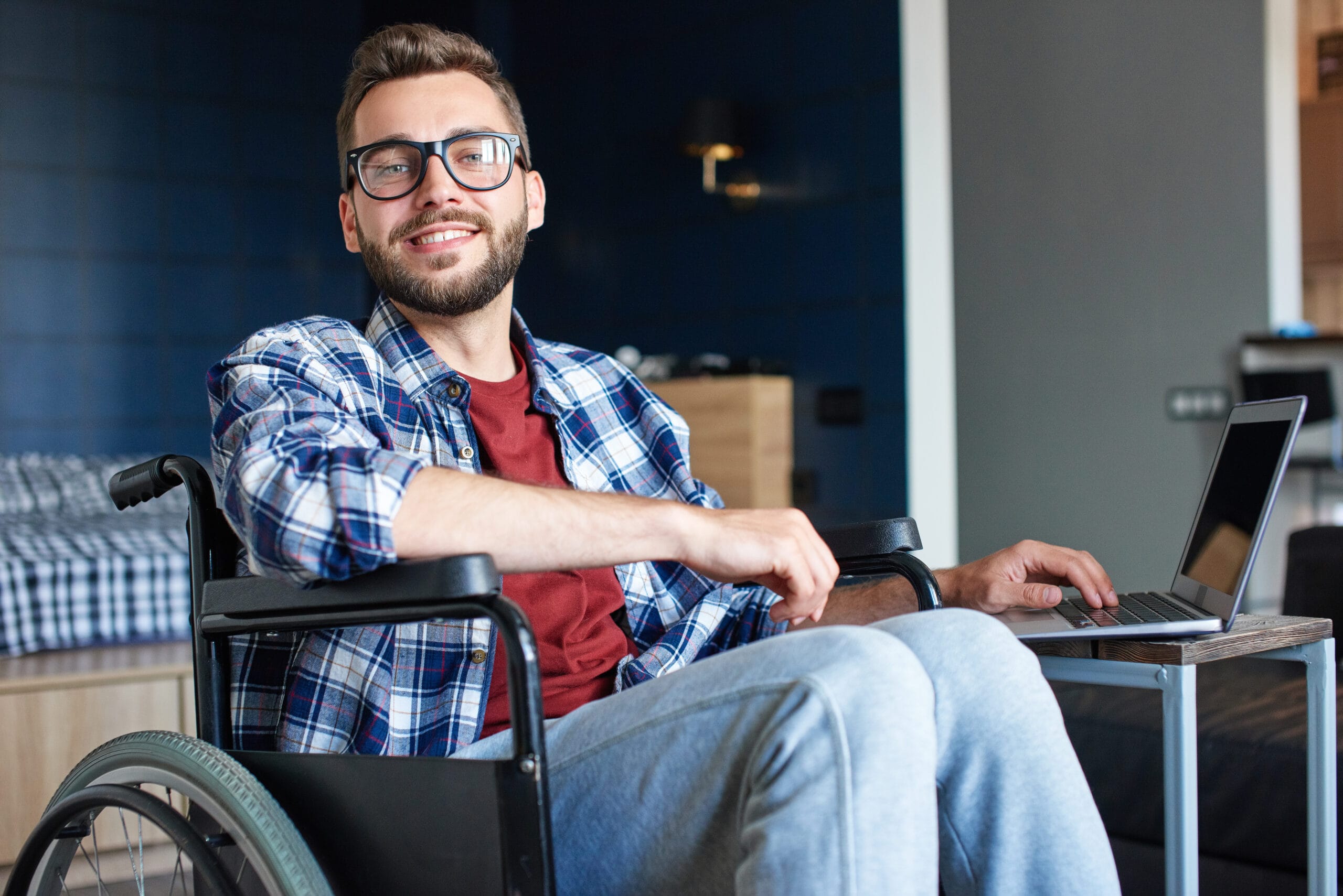 Young man with beard in wheelchair, working on laptop from home, answering call. He is wearing plaid blue shirt, sitting at his kitchen table.