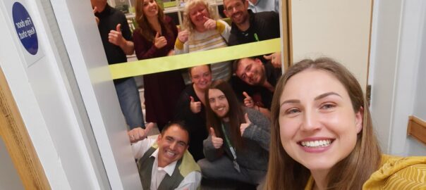 A group photo of the full Direct Access Team at the grand opening of the office. A yellow banner ready to be cut is stretched across the door.