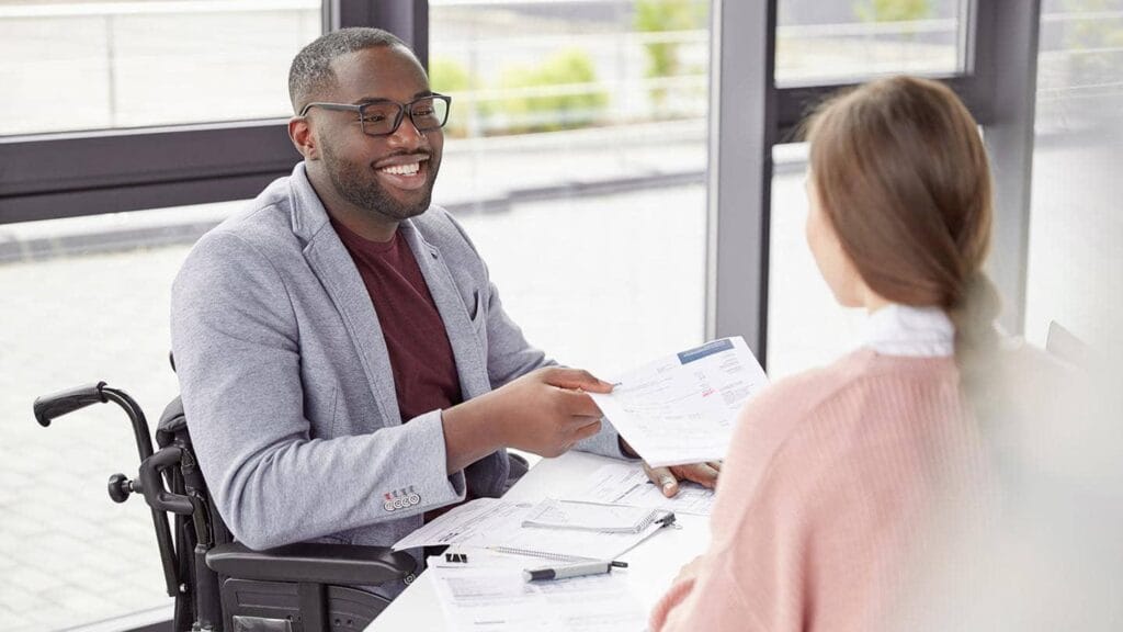 A man in a wheelchair hands a document to a female colleague across a desk in an office.